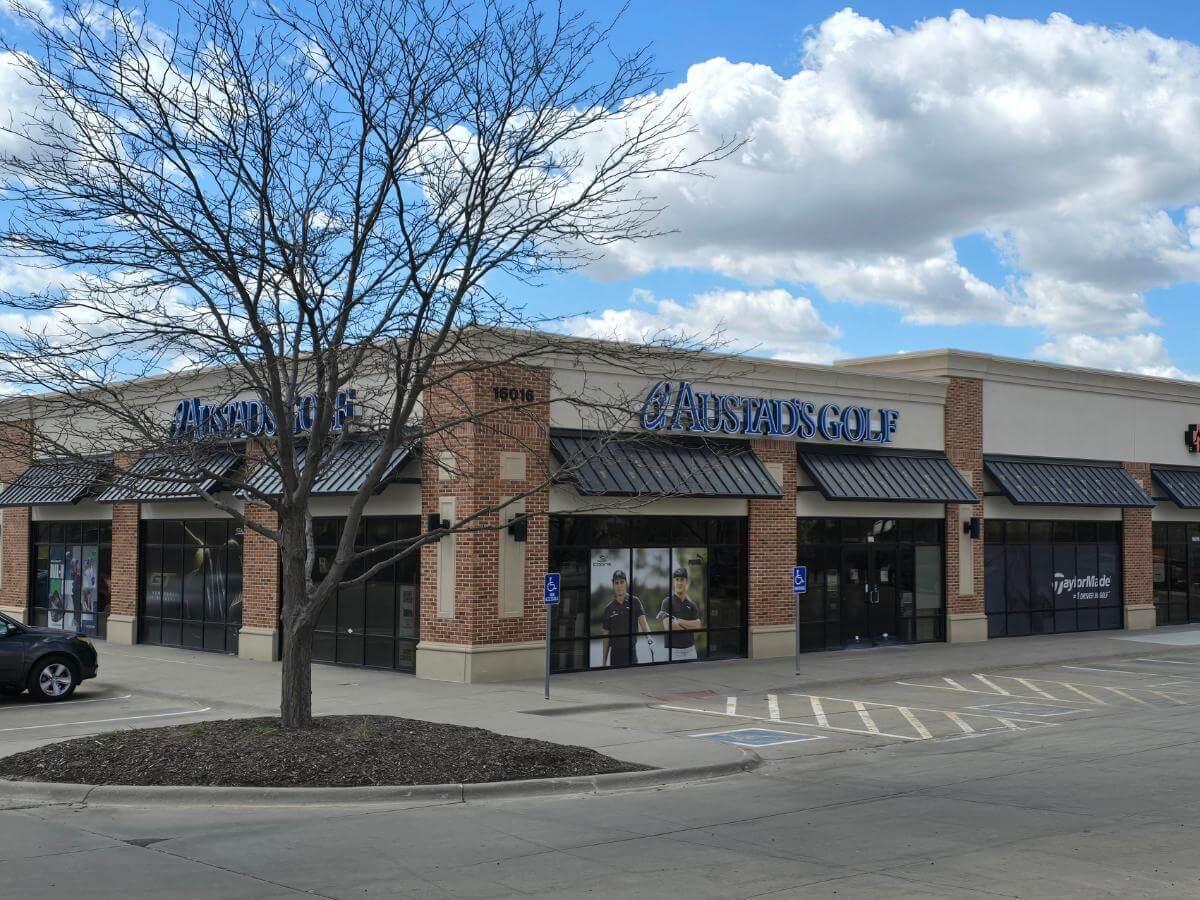 The exterior of Austad's Golf in West Maple- a smaller building that looks to be a part of a plaza. It's a corner store with cream colored concrete walls and red bricked columns.