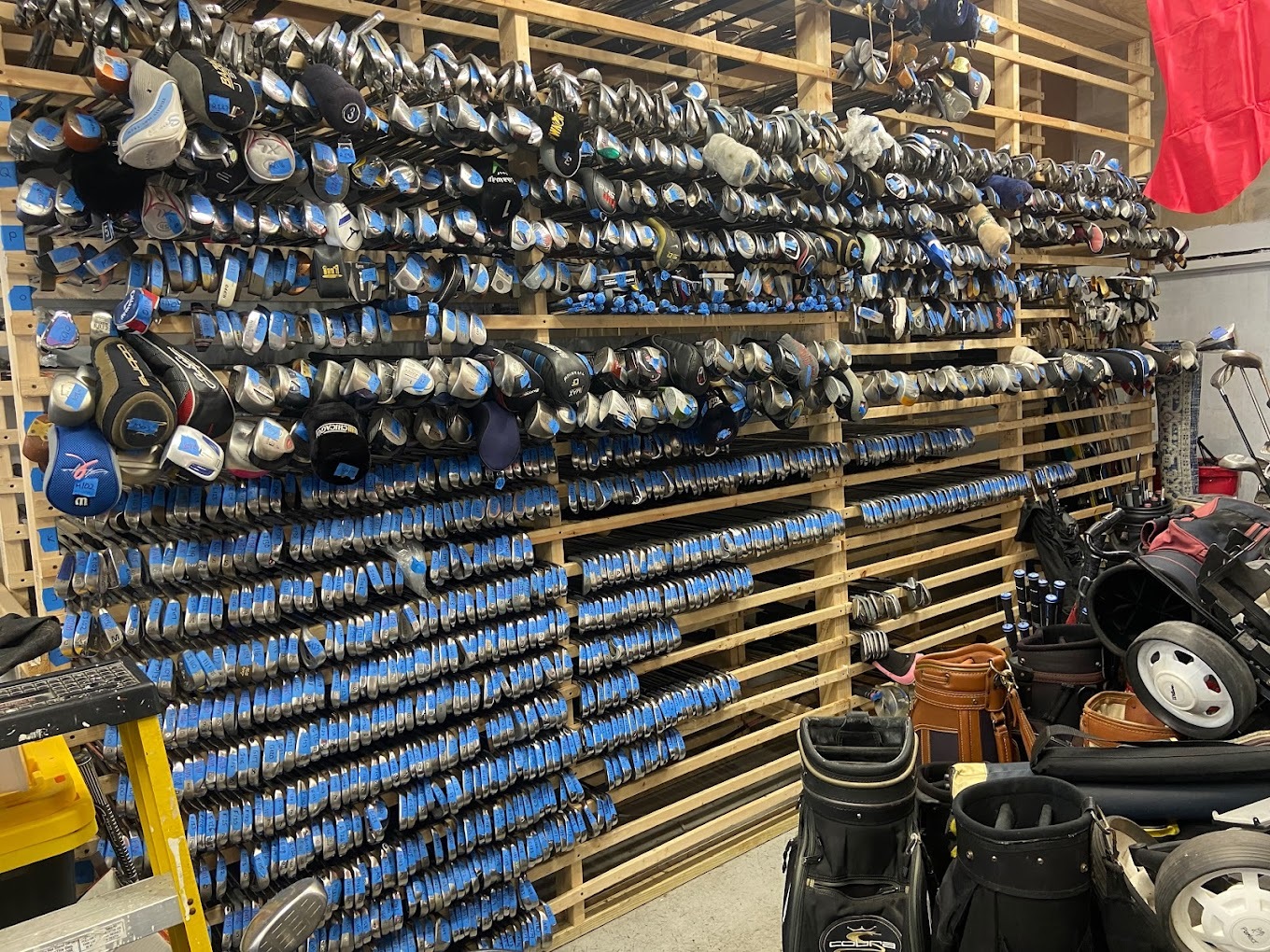 A tall and wide wooden rack holding hundreds of golf clubs with golf bags on the floor beside it.