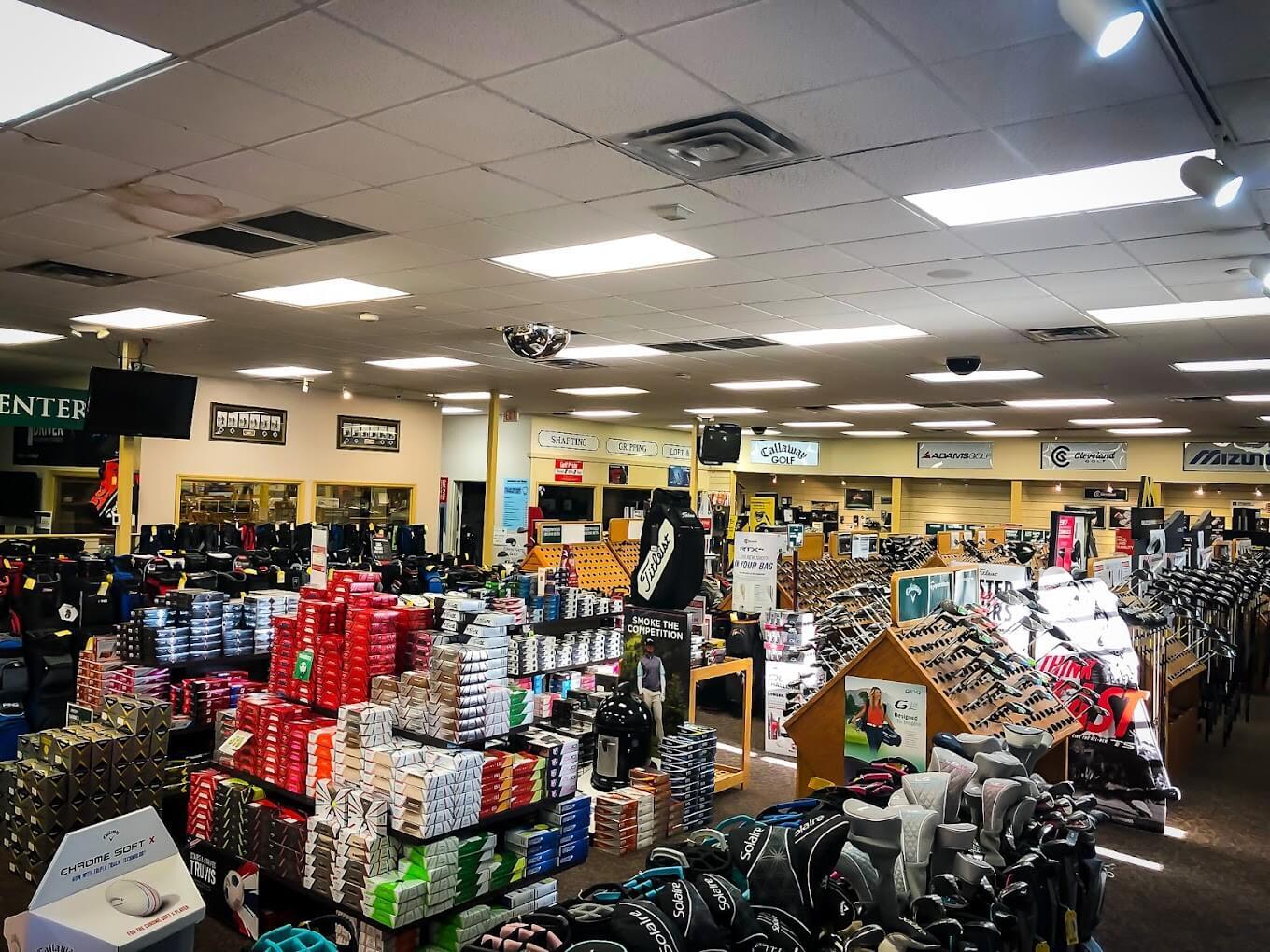 The inside of Edwin Watts Golf features stacked boxes of balls, clubs in racks, bags, and more on the store floor on a sunny afternoon.