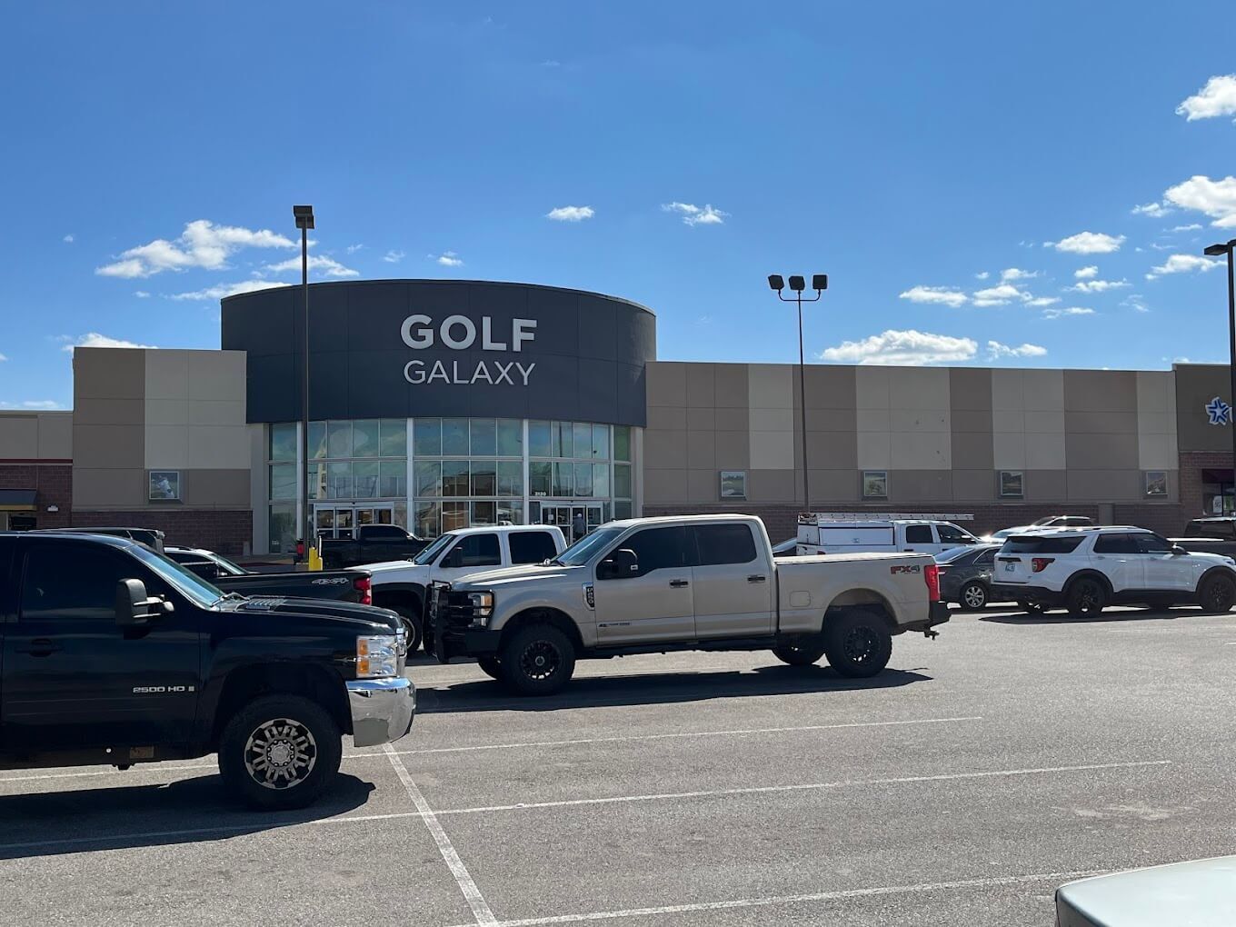 The exterior of Golf Galaxy on a sunny day with small white clouds in the sky. The photo is taken from 100 feet back in the parking lot which features trucks and cars.