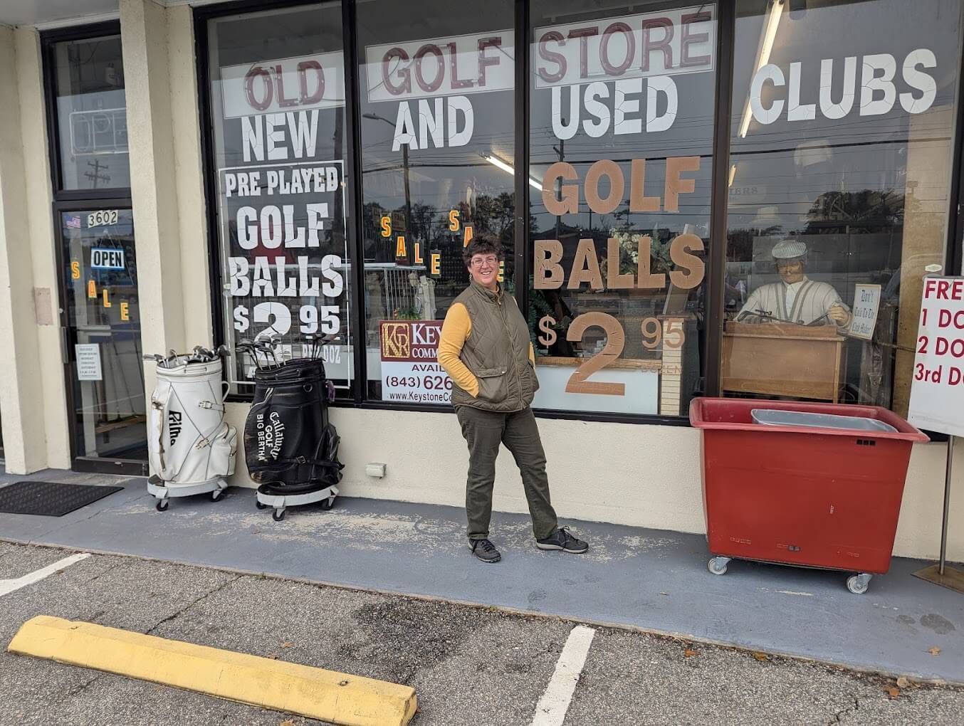 The exterior of the Old Golf Store featuring big vertical windows with lettering advertising their "new and used clubs" along with $2.95 golf balls. There is a short-haired middle-aged white woman standing in front of the windows smiling. She is wearing glasses, a light brown vest, a yellow long-sleeved shirt, and dark khakis.