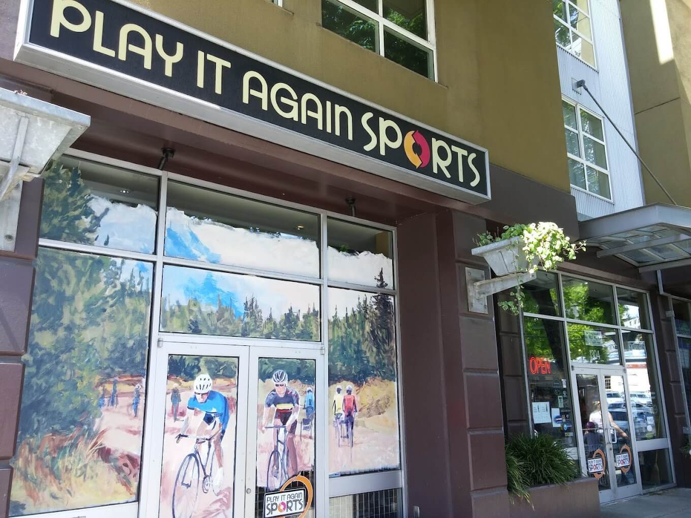 The front exterior of Play It Again Sports - Seattle which features a small window front with an artists' depiction of bikers on a trail on a sunny afternoon.