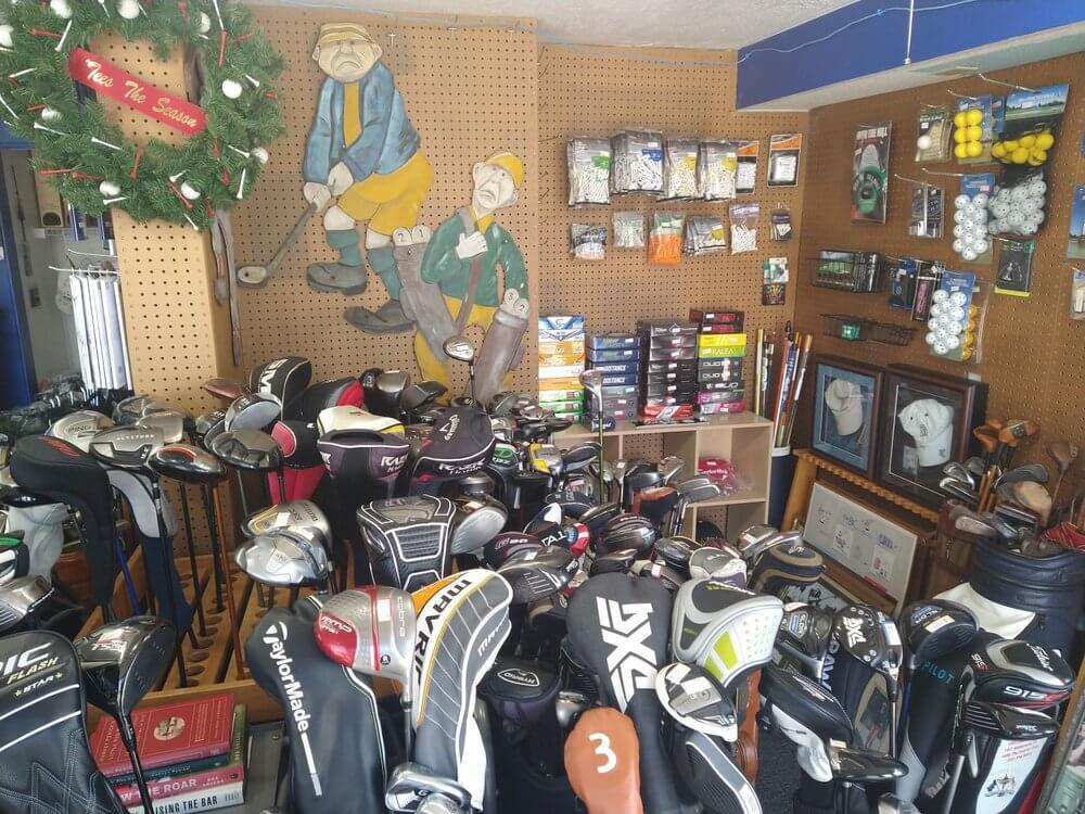 A corner of the interior of Pinecraft Golf Shop that's full of inventory. There's multiple golf bags holding used golf clubs along with balls hung on the wall and on display shelves, along with an artist rendition of to cartoon golfers hanging on the wall.