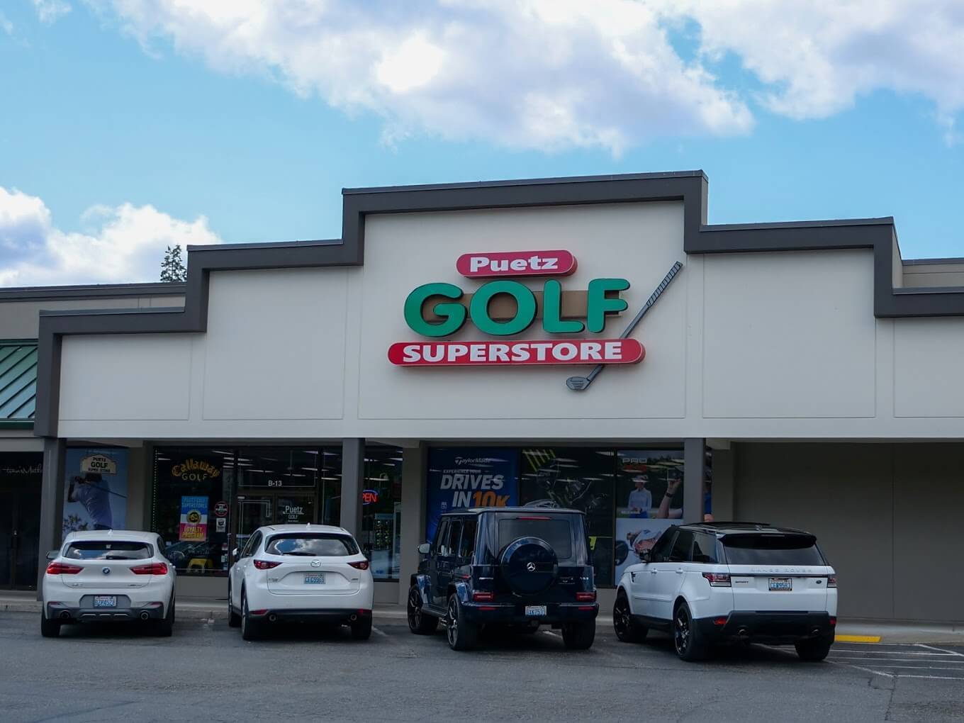 The front exterior of Puetz Golf Bellevue Superstore. It's located in a strip-mall with beige and grey paint and a big green and red sign displaying the store's name. There are four cars parked in front of it and there are advertisements in the windows.