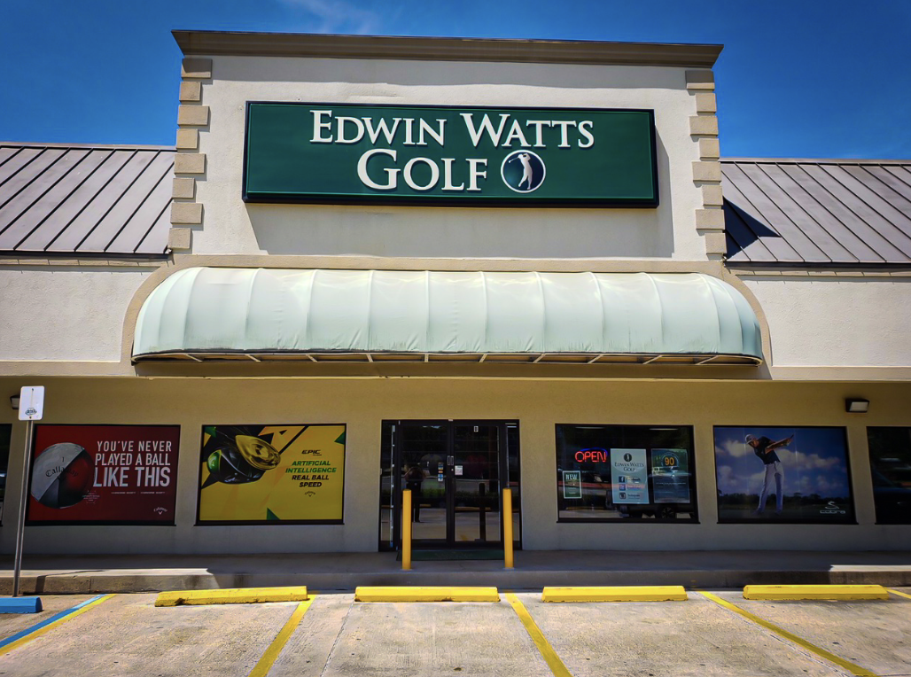 The outside of Edwin Watts Golf in NOLA on a sunny day. The building is beige and big and features advertisements on the windows for golf products.