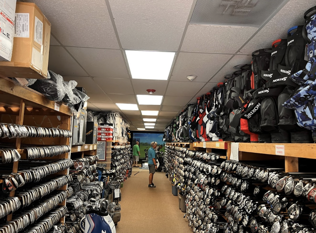 The interior of Golf Trader showing wooden shelves of hundreds of used golf clubs and bags for sale with three people shopping in the background.