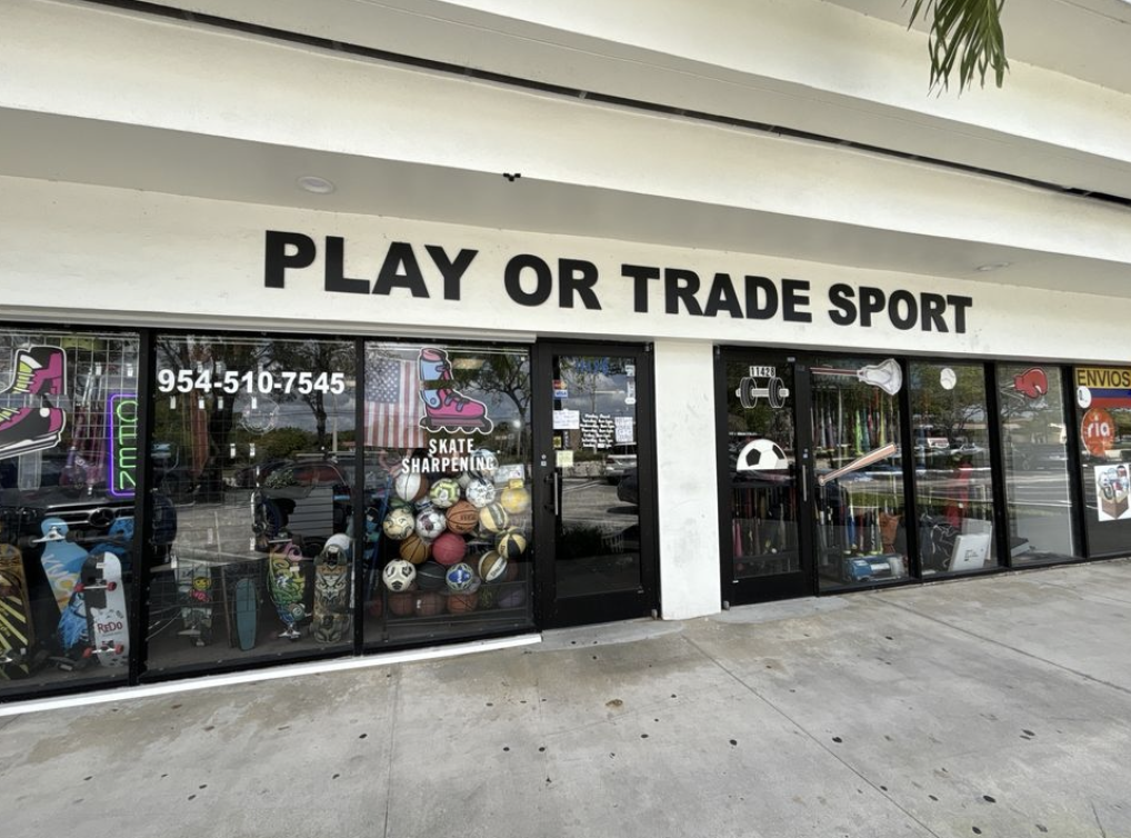 The exterior of Play or Trade Sports. It's a white building with ground to top windows featuring a ton of stacked sporting equipment.