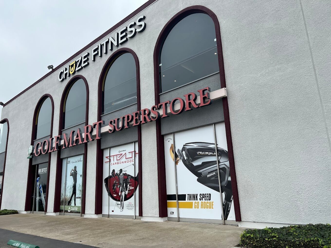 The exterior of Golf Mart Superstore is a light gray building with floor to ceiling arch windows that have big pictures of gold club brands and golf players displayed on a cloudy day.
